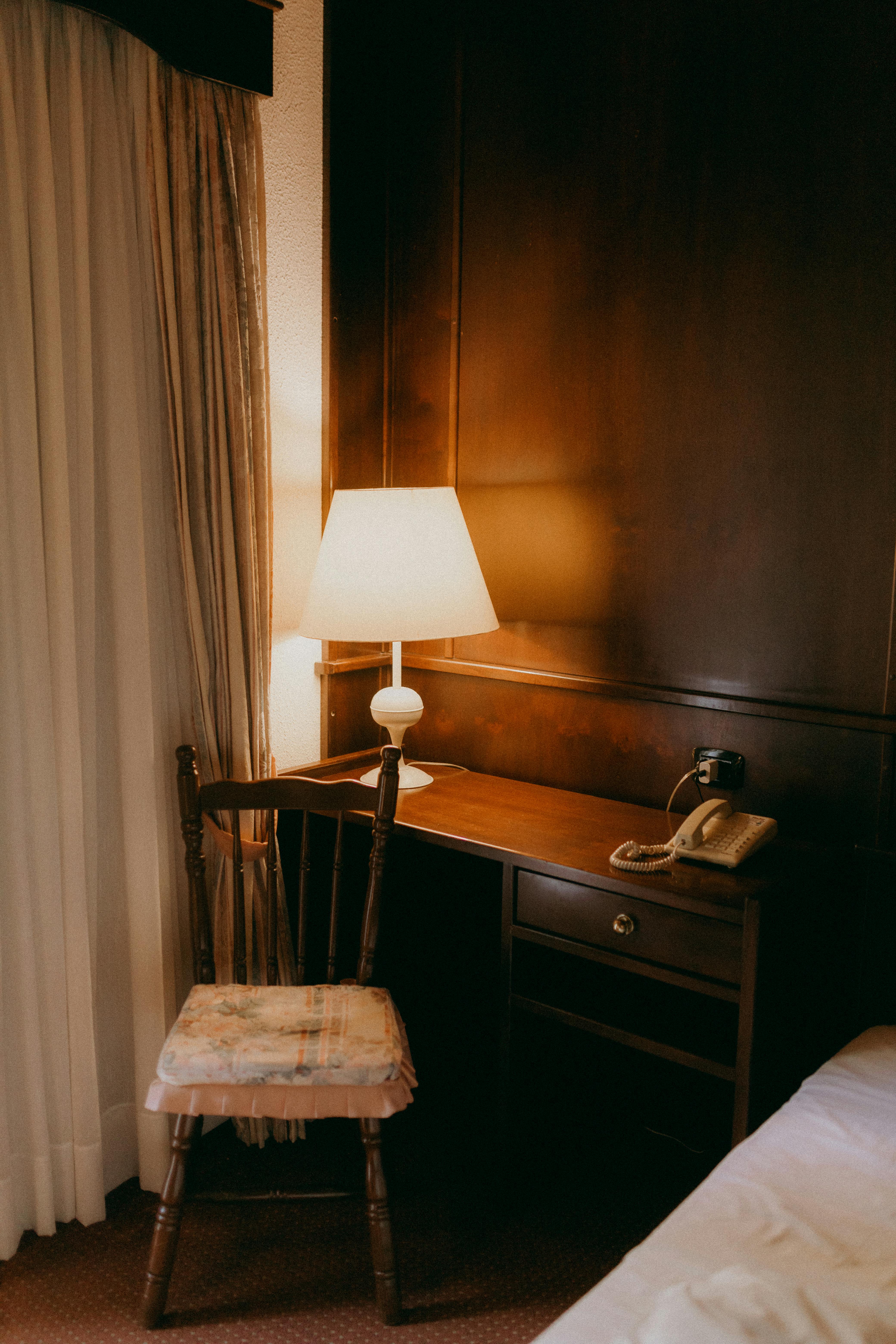 Warmly lit hotel room corner with a desk, chair, and lamp for a cozy ambiance.